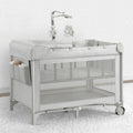 A babyGap Deluxe Play Yard with a infant inside, featuring mesh sides, a white frame, and wheels for mobility.