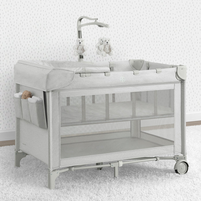 A babyGap Deluxe Play Yard with a infant inside, featuring mesh sides, a white frame, and wheels for mobility.