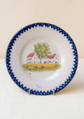 A plate with hand painted house motif and blue patterned rim.