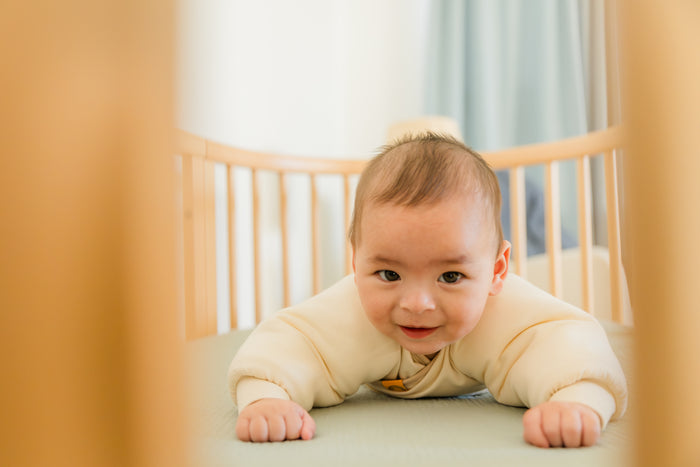 Baby in a crib wearing a coconut cream Jacky swaddle transition jacket, on belly looking at the camera.