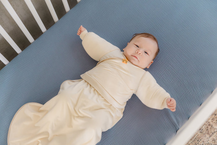 baby wearing coconut cream Jacky swaddle transition jacket layered over coconut cream sleep bag laying on back in a crib