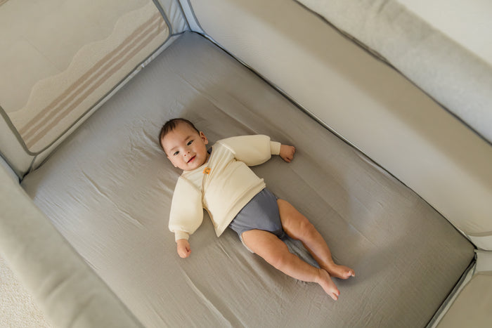baby wearing coconut cream Jacky swaddle transition jacket layered over gray onesie laying on back in a portable crib