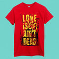 A classic-fit red '68 Gildan T-shirt in cotton polyester features bold yellow and black "LOVE ISNT DEAD" text on a blue textured background, with "ISNT" formed by red marks striking out "IS.