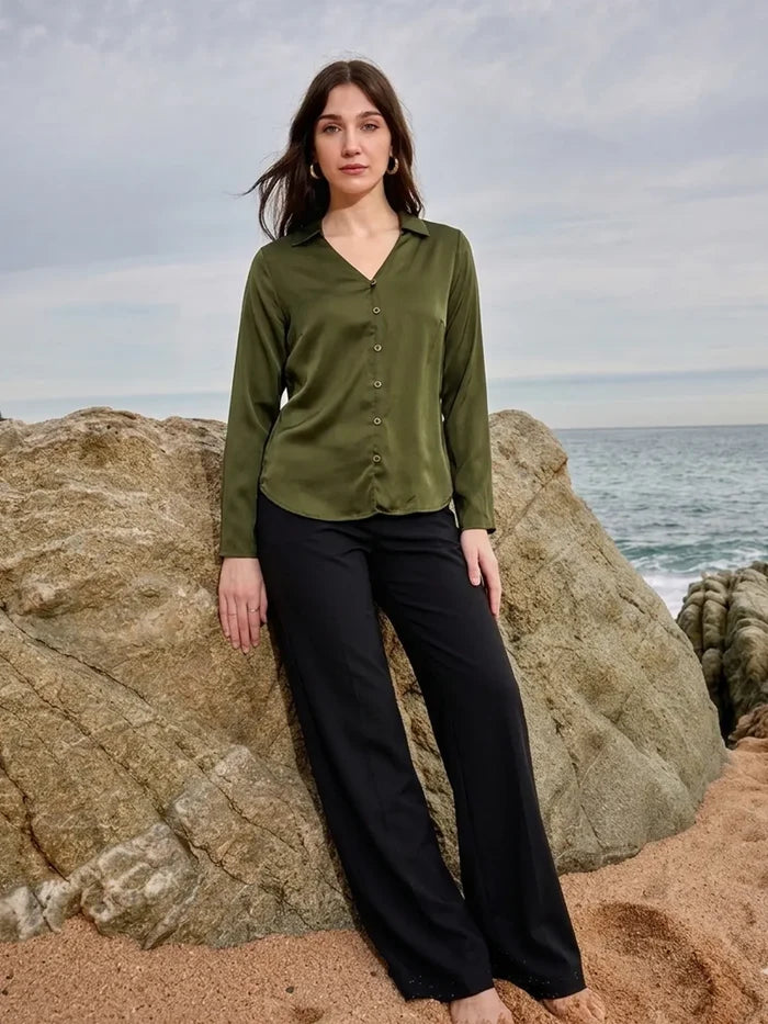 Woman wearing a green shirt and black pants standing on rocky terrain by the ocean.