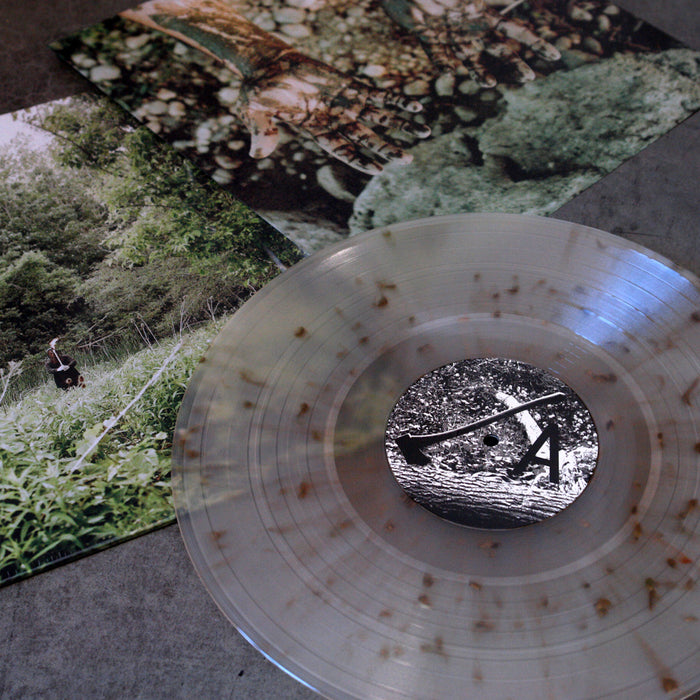 A clear Exalt Palmcarver vinyl record with brown speckles rests on a gray surface, partly covering its "Death Is A Road" album cover, which displays nature-themed photos of greenery, trees, and a person in tall grass.